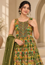 A woman models a sleeveless green anarkali-style dress with a multicolor zigzag print and ornate gold embroidery on the bodice, paired with a matching olive green dupatta in a warm, arched interior setting.