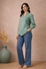 Seafoam green asymmetrical blouse side angle, long sleeves and curved hem, paired with blue jeans and block heels.