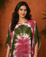Close-up portrait of a South Asian female model wearing an olive and maroon tie-dye kaftan, standing against a warm terracotta textured wall with soft studio lighting and subtle greenery in the background.