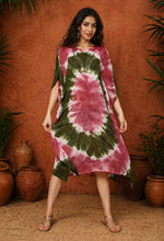 Front-facing full-length studio photograph of a South Asian female model wearing an olive and maroon tie-dye kaftan, standing against a warm terracotta textured wall with decorative clay pots and green plants, captured in professional fashion catalog lighting.