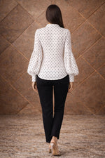 Woman wearing a white blouse with black polka dots and black pants against a brown textured wall.