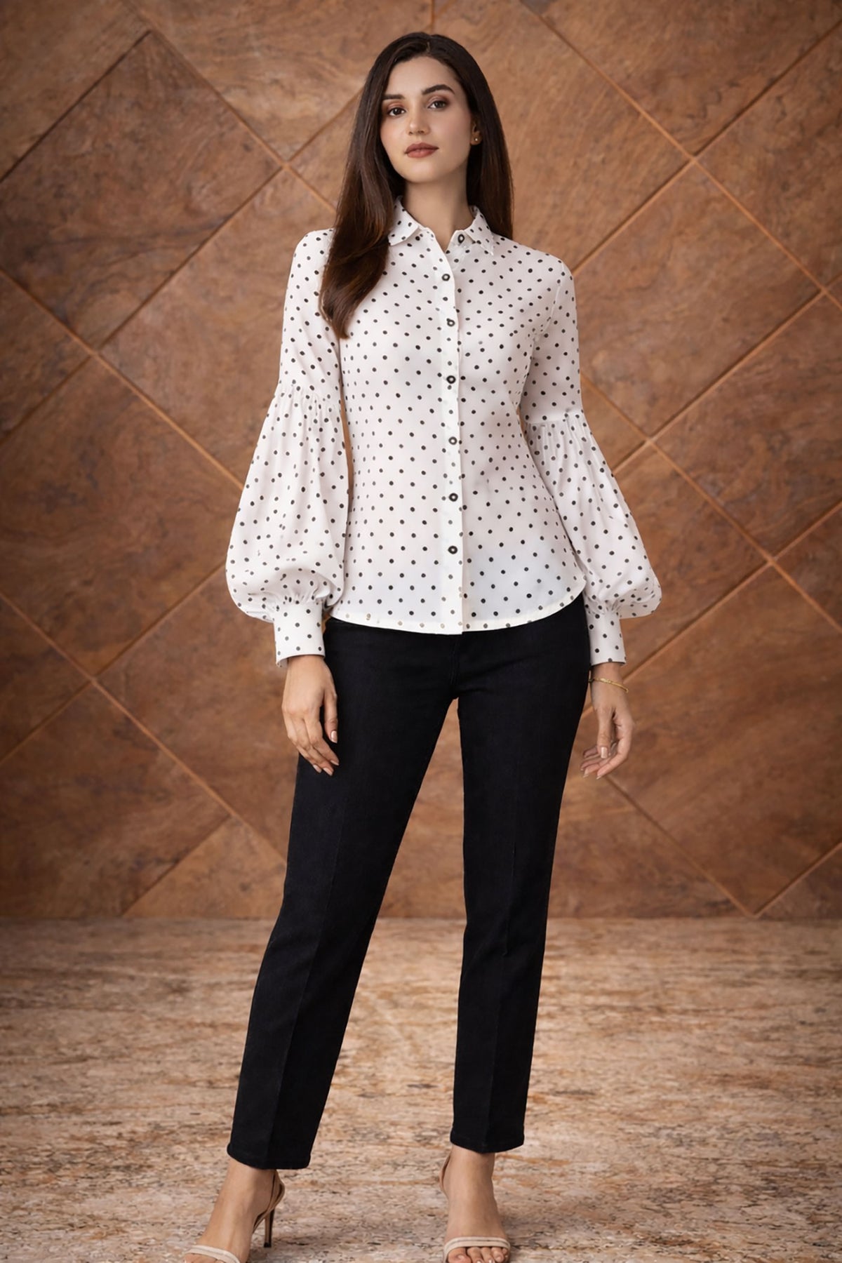 Woman wearing a white blouse with black polka dots and black pants against a brown textured wall.