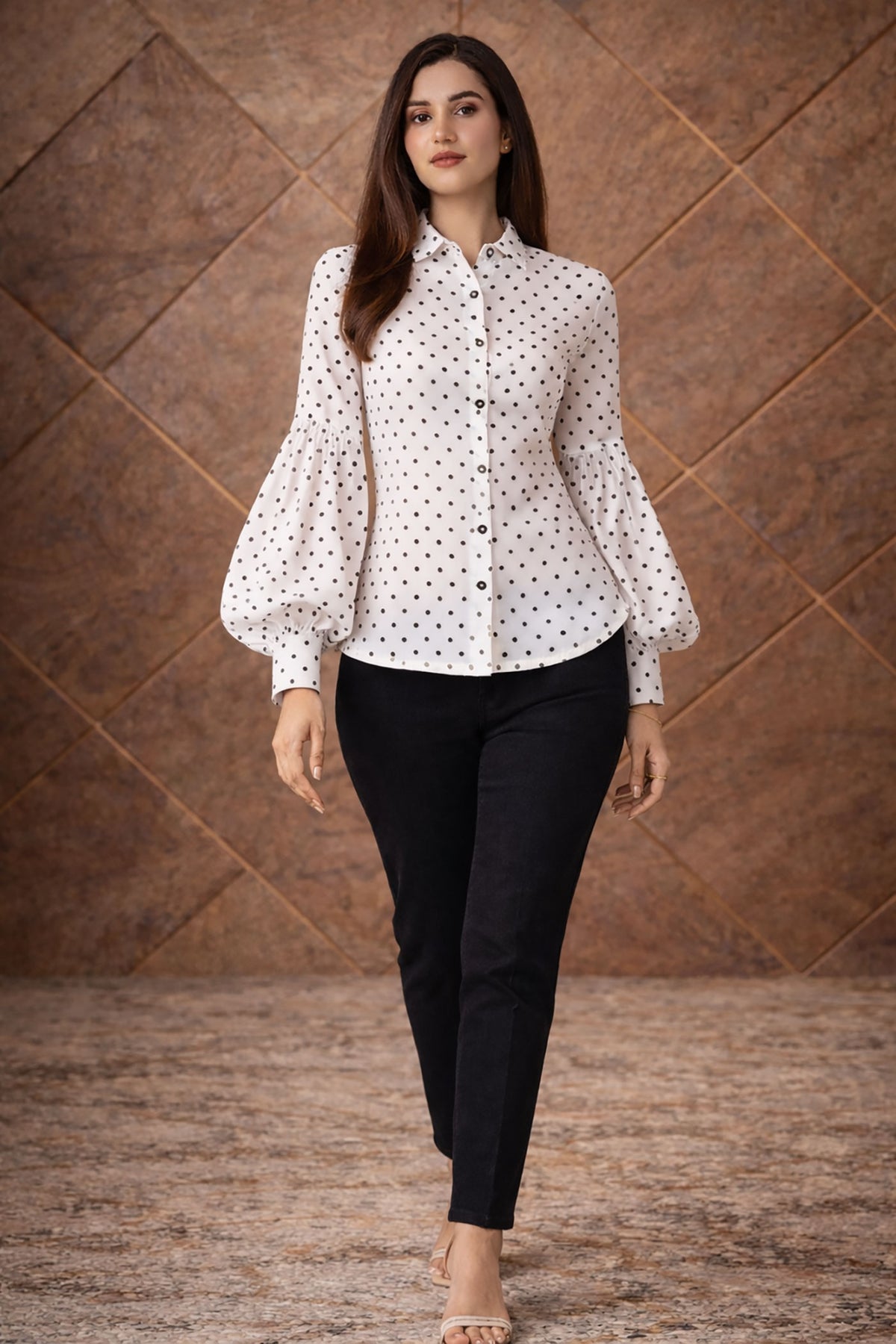 Woman wearing a white blouse with black polka dots and black pants against a brown textured wall.