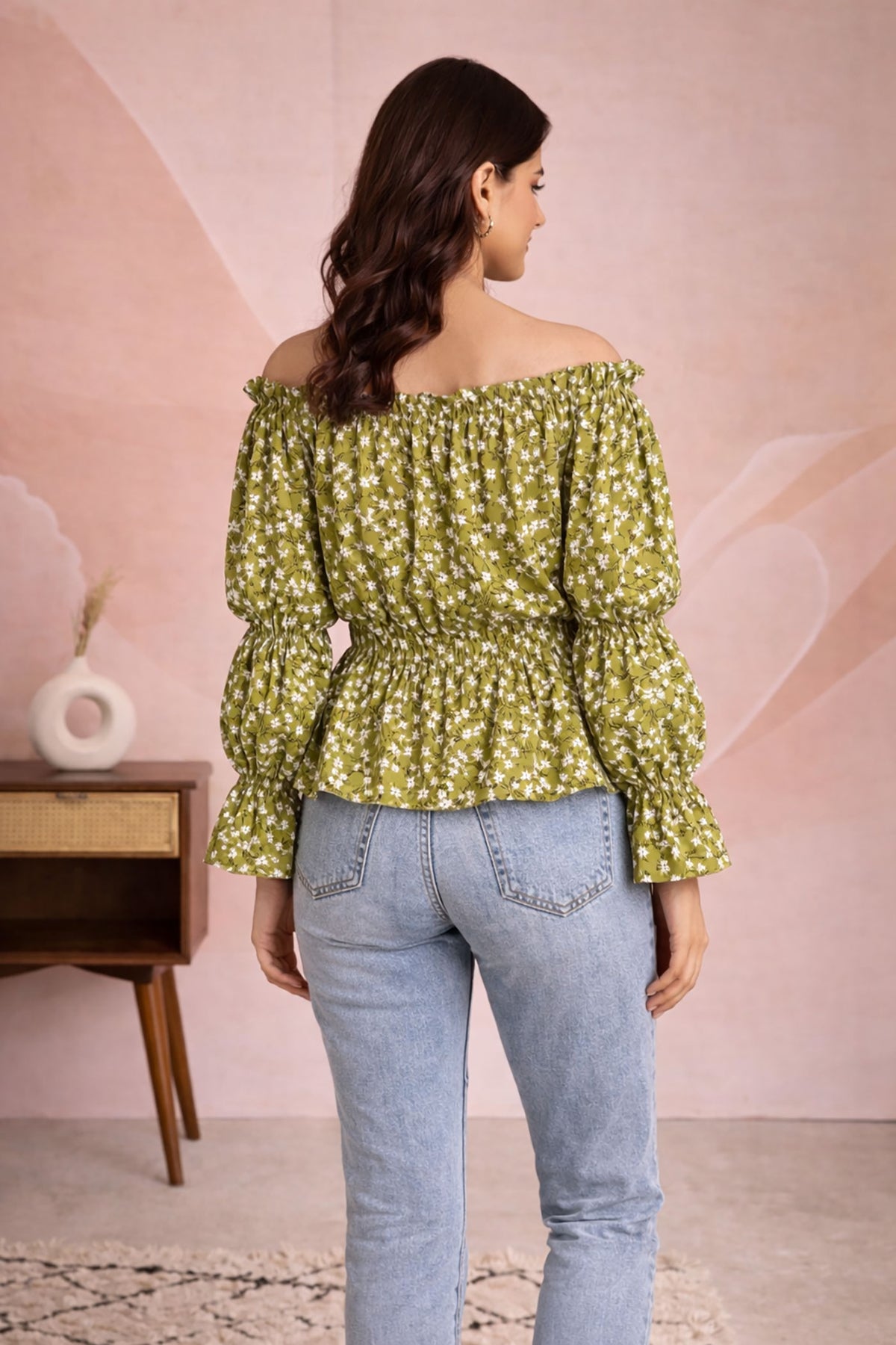 Woman wearing a green floral off-shoulder top and light blue jeans against a pink wall.