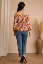 Woman wearing an off-shoulder top with abstract pattern and blue jeans, standing against a beige wall.