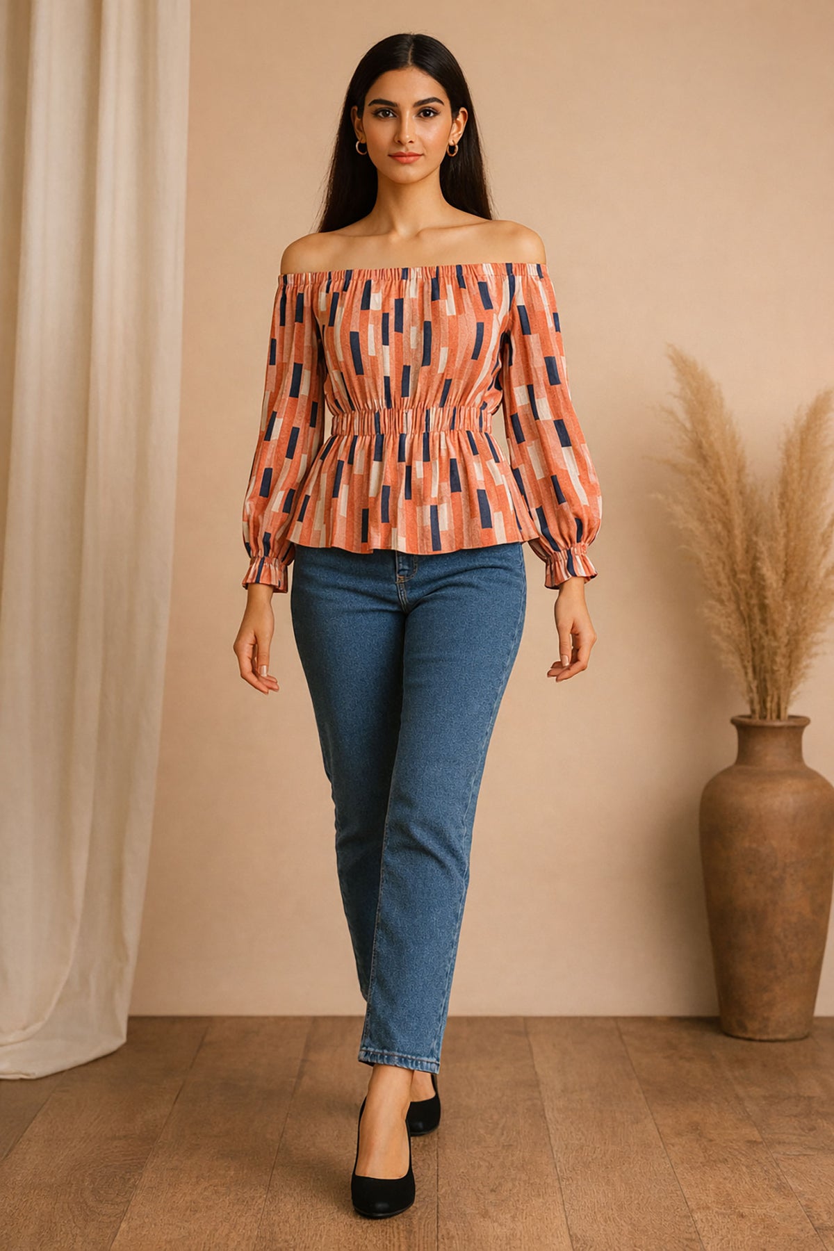 Woman wearing an off-shoulder patterned top and blue jeans standing against a beige wall.