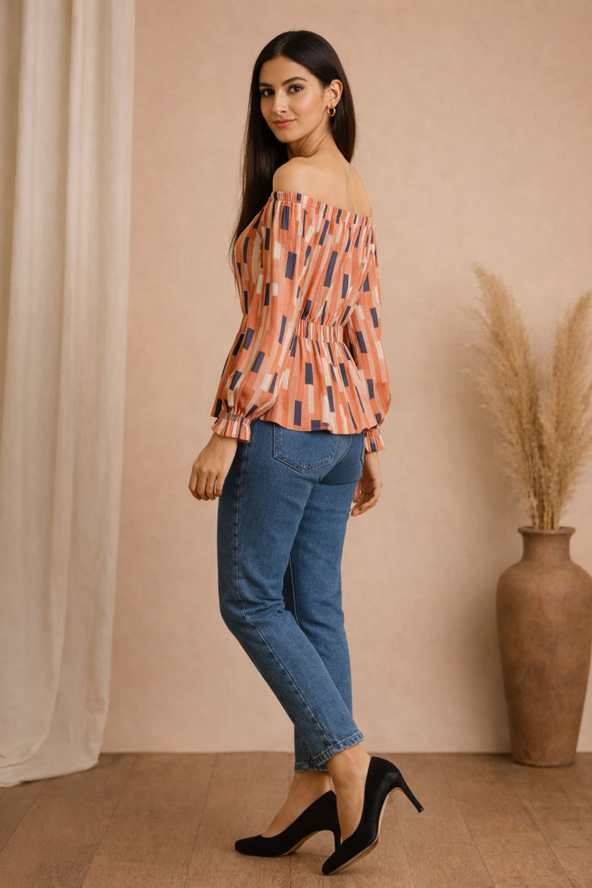 Woman wearing a patterned blouse and blue jeans standing against a beige wall with a vase of dried plants.