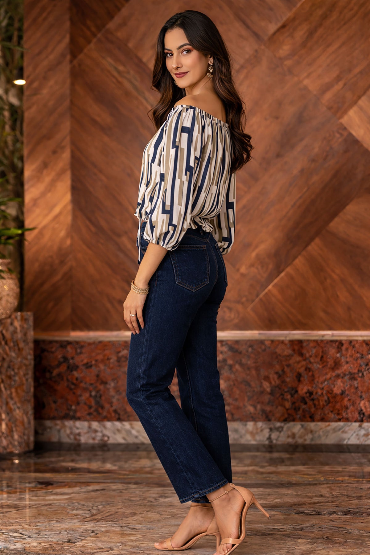 Woman wearing a patterned blouse and dark jeans against a wooden wall.