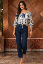Woman wearing a patterned top and dark jeans standing against a wooden wall.