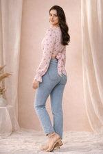 Woman wearing a pink floral blouse and blue jeans standing against a beige curtain backdrop.