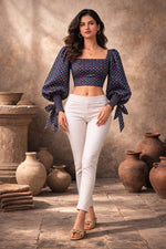 Woman wearing a dark blue patterned top and white pants standing in front of rustic pots.