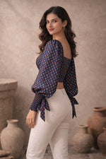 Woman wearing a blue patterned top and white pants standing in front of rustic vases.