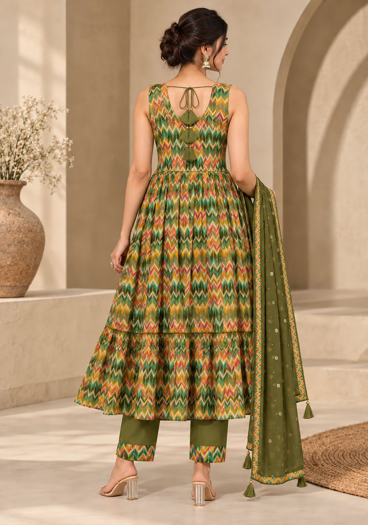 A woman is shown from the back wearing a sleeveless olive green and orange geometric print salwar kameez set with matching pants and a dupatta, in a warm beige architectural setting.