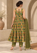 A woman is shown from the back wearing a sleeveless olive green and orange geometric print salwar kameez set with matching pants and a dupatta, in a warm beige architectural setting.