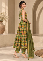 A woman models a green traditional Indian outfit with a multicolor geometric patterned kurta, matching pants, and a solid green dupatta in a warm, beige-toned architectural setting.