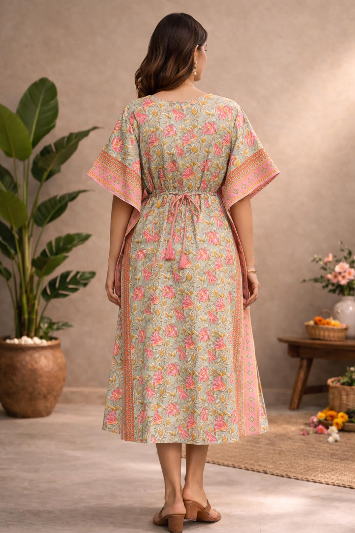Back view of woman wearing pastel green and pink floral cotton kaftan dress with tie-up waist and border detailing in studio setting