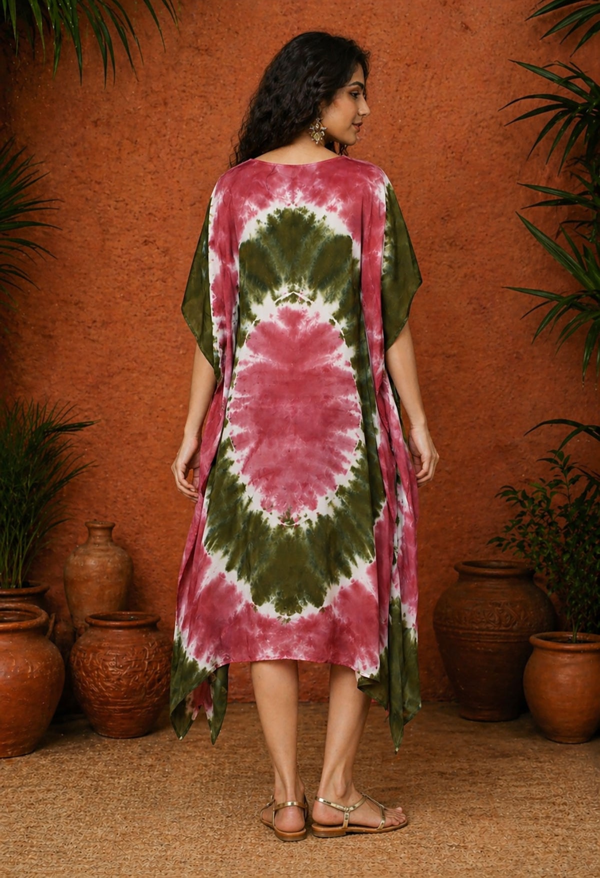Back view of a South Asian female model wearing an olive and maroon tie-dye kaftan, standing against a warm terracotta textured wall with clay pots and greenery in soft studio lighting.