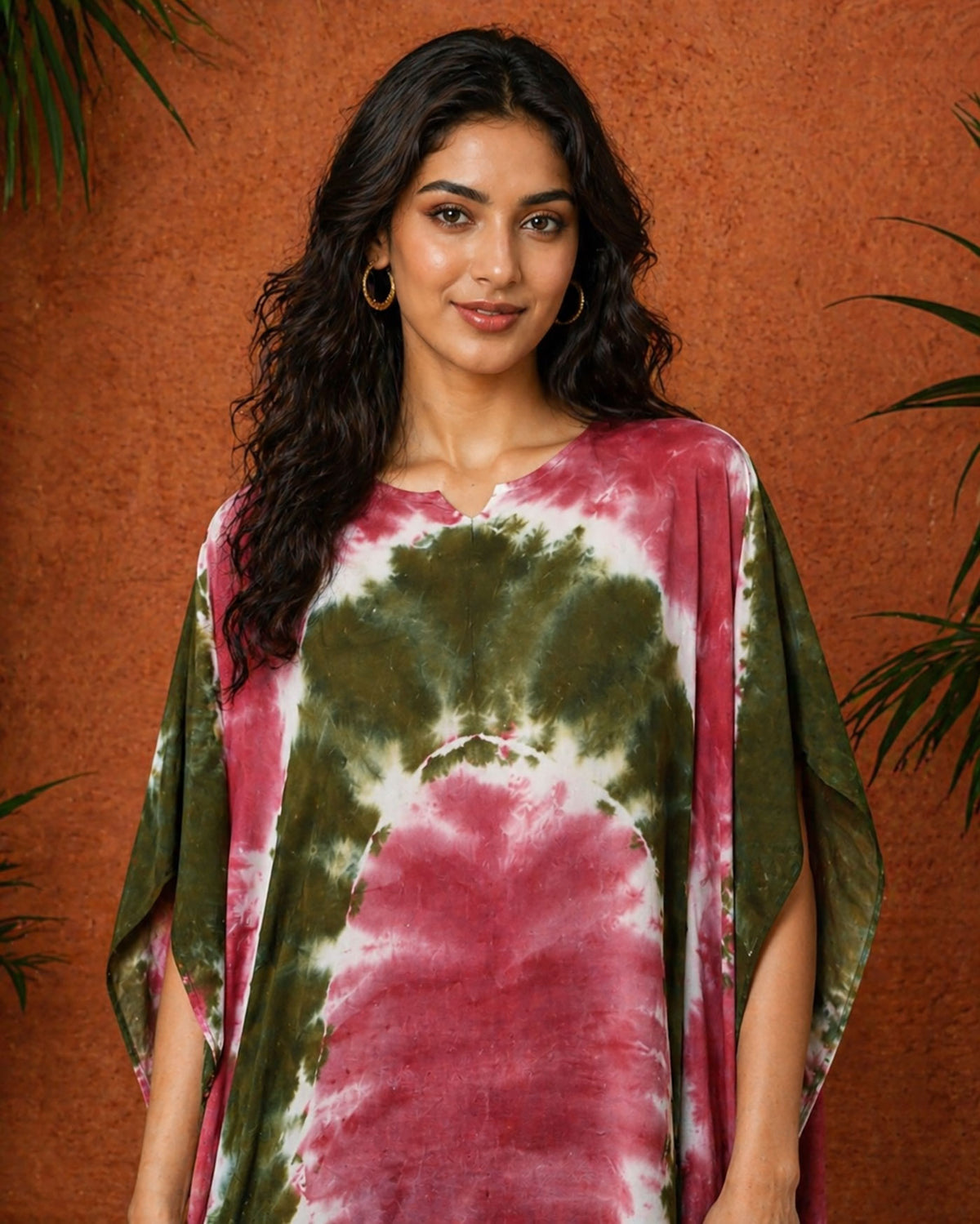 Close-up portrait of a South Asian female model wearing an olive and maroon tie-dye kaftan, standing against a warm terracotta textured wall with soft studio lighting and subtle greenery in the background.