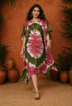 Full-length image of a South Asian female model walking toward the camera wearing an olive and maroon tie-dye kaftan, set against a warm terracotta textured wall with clay pots and greenery in soft studio lighting.