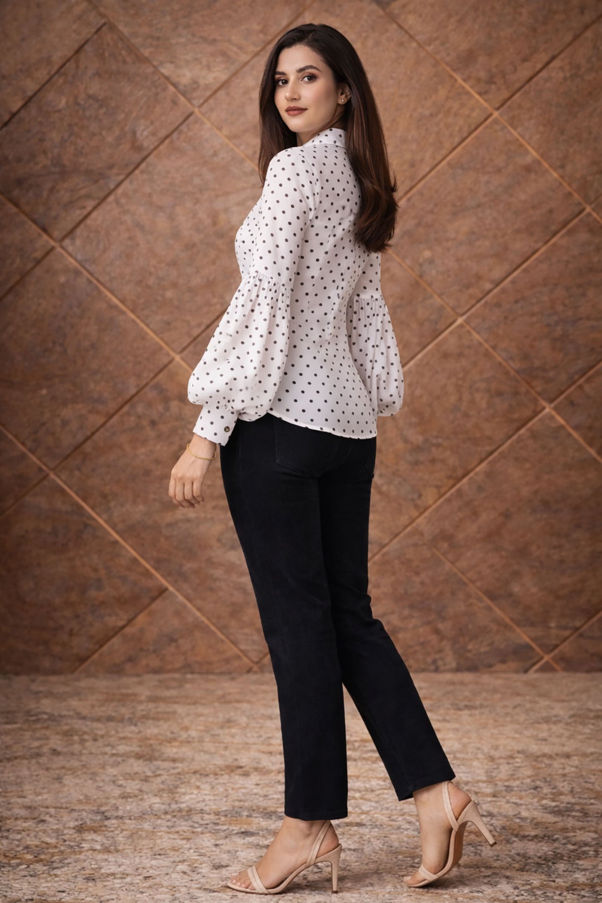 Woman wearing a white polka dot blouse and black pants against a brown textured wall.