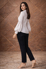 Woman wearing a white polka dot blouse and black pants against a brown textured wall.