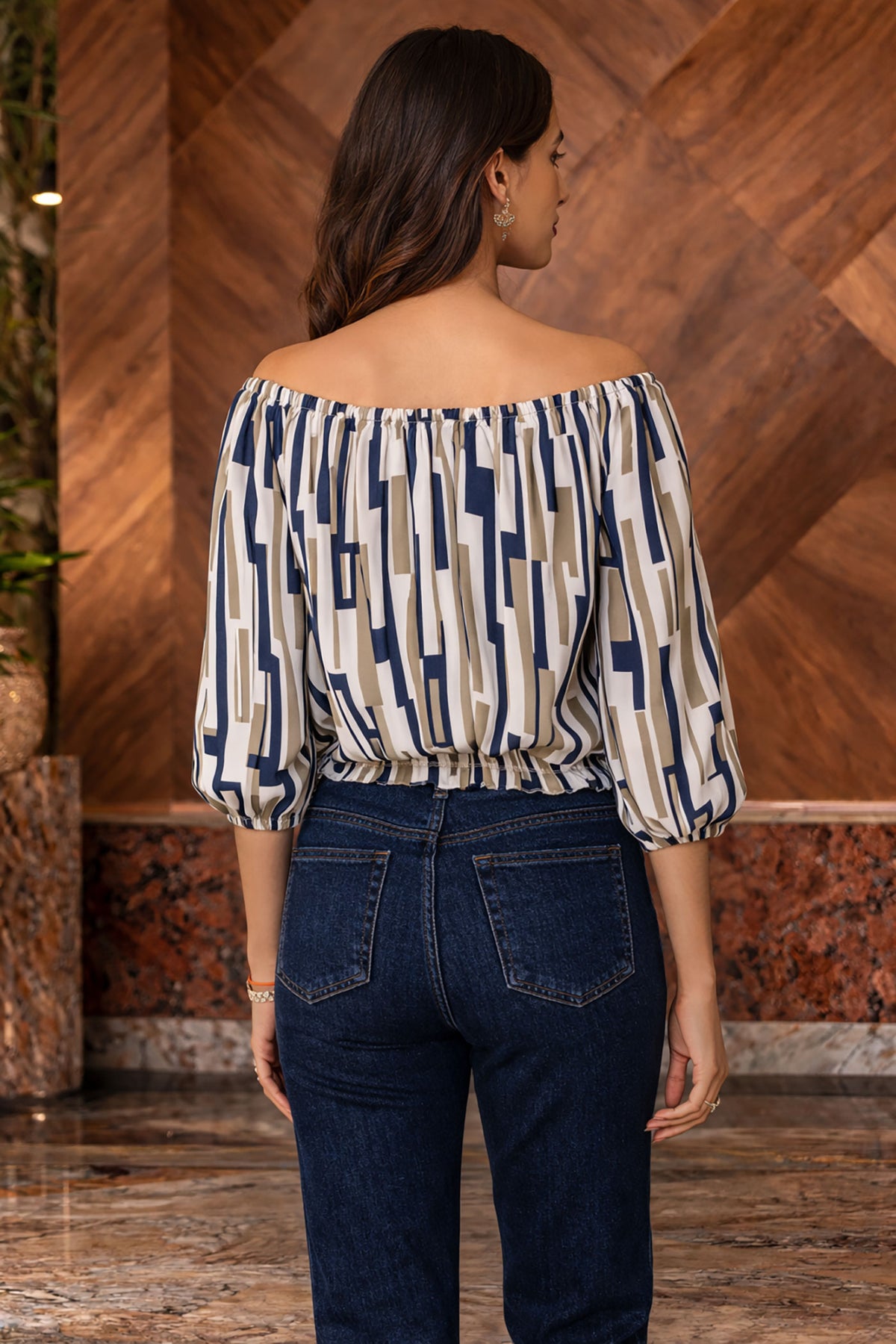 Woman wearing a patterned top and dark jeans against a wooden wall.