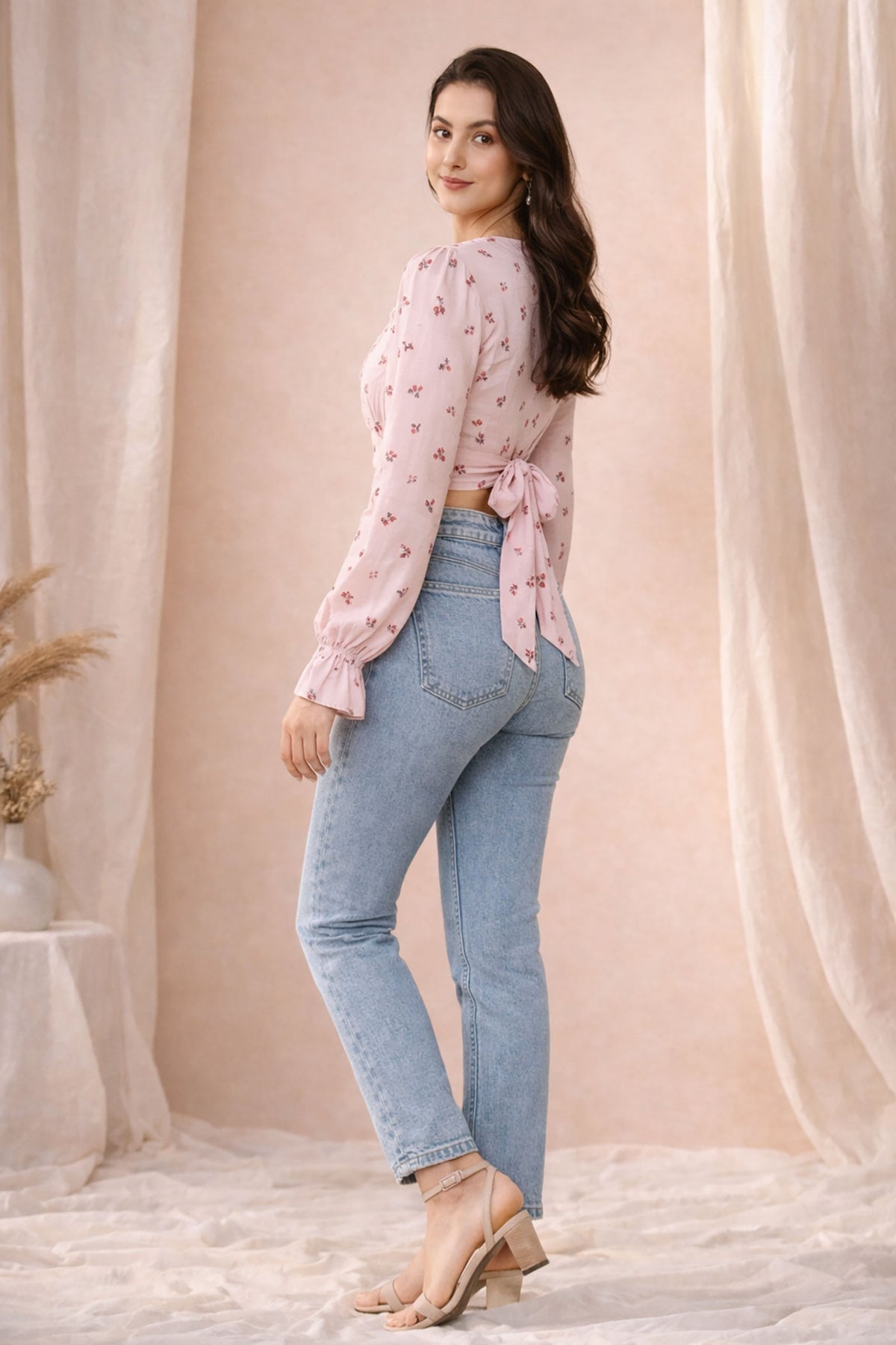 Woman wearing a pink floral blouse and blue jeans standing against a beige curtain backdrop.